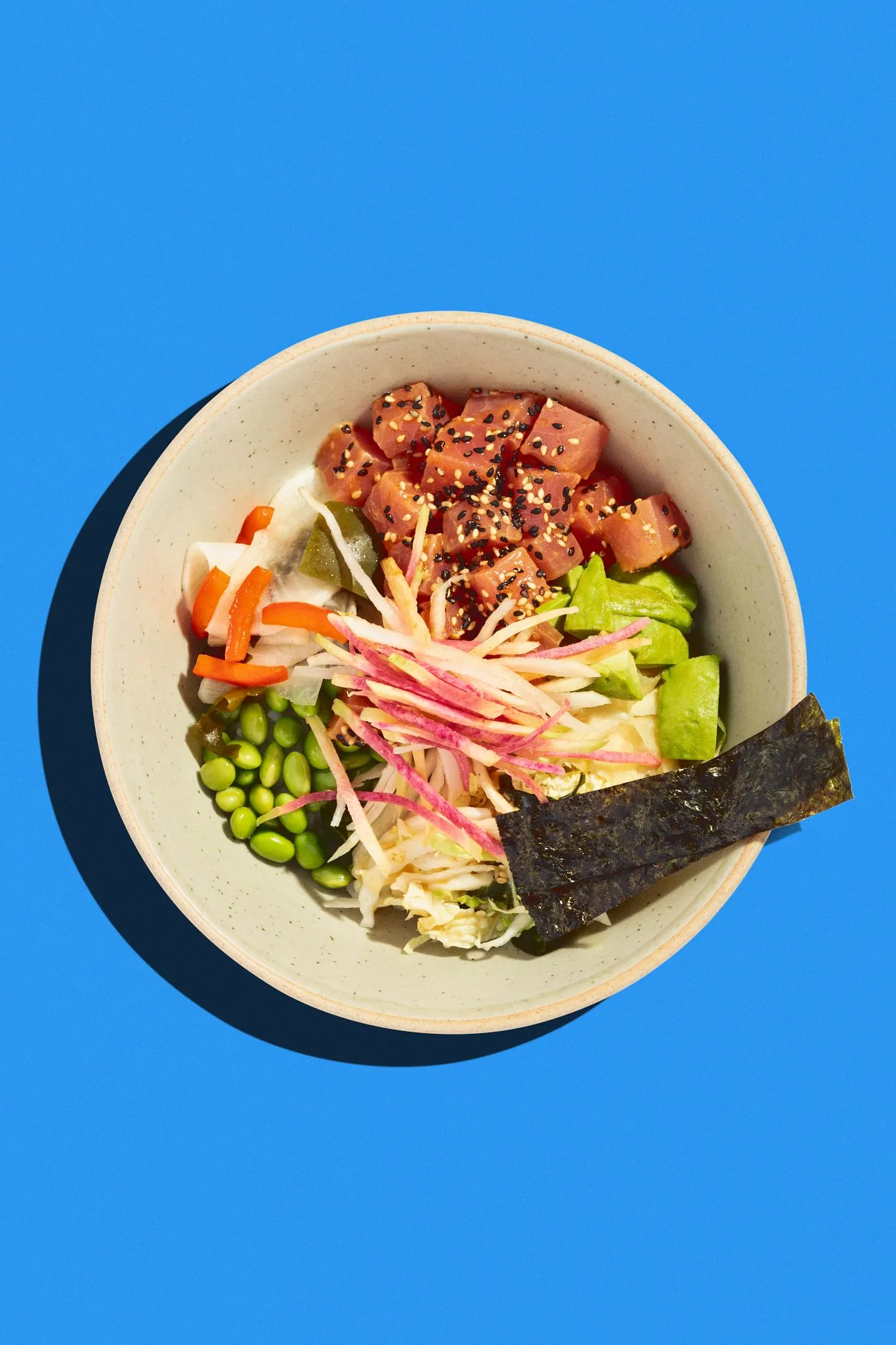 Overhead shot of a poke bowl with tuna, vegetables, and seaweed on a bright blue background.