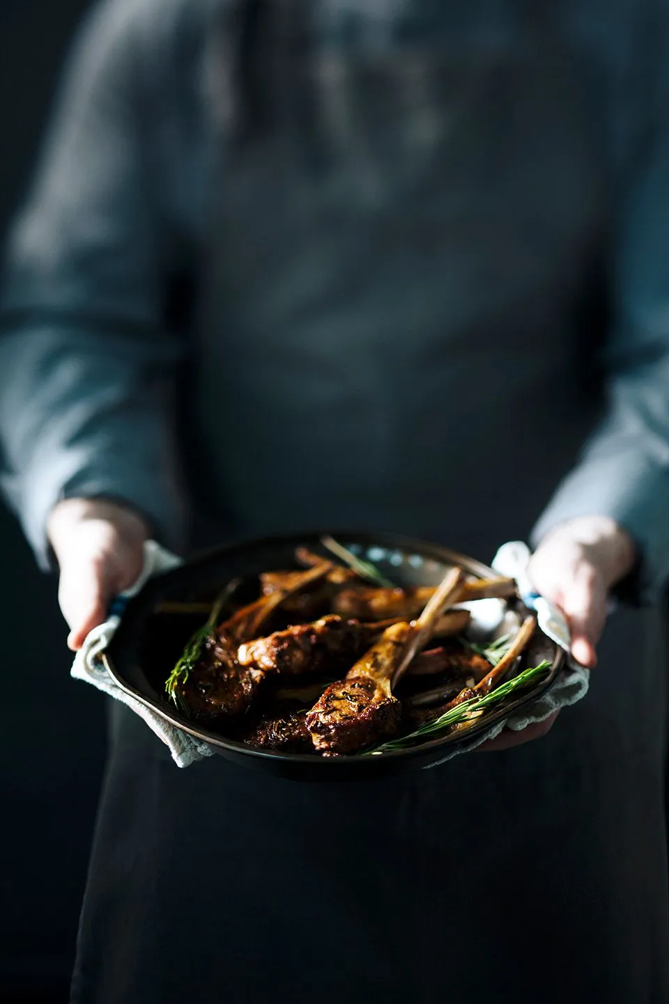 A person in a gray apron and shirt holds a dark pan of cooked lamb chops garnished with rosemary, set on a white cloth.