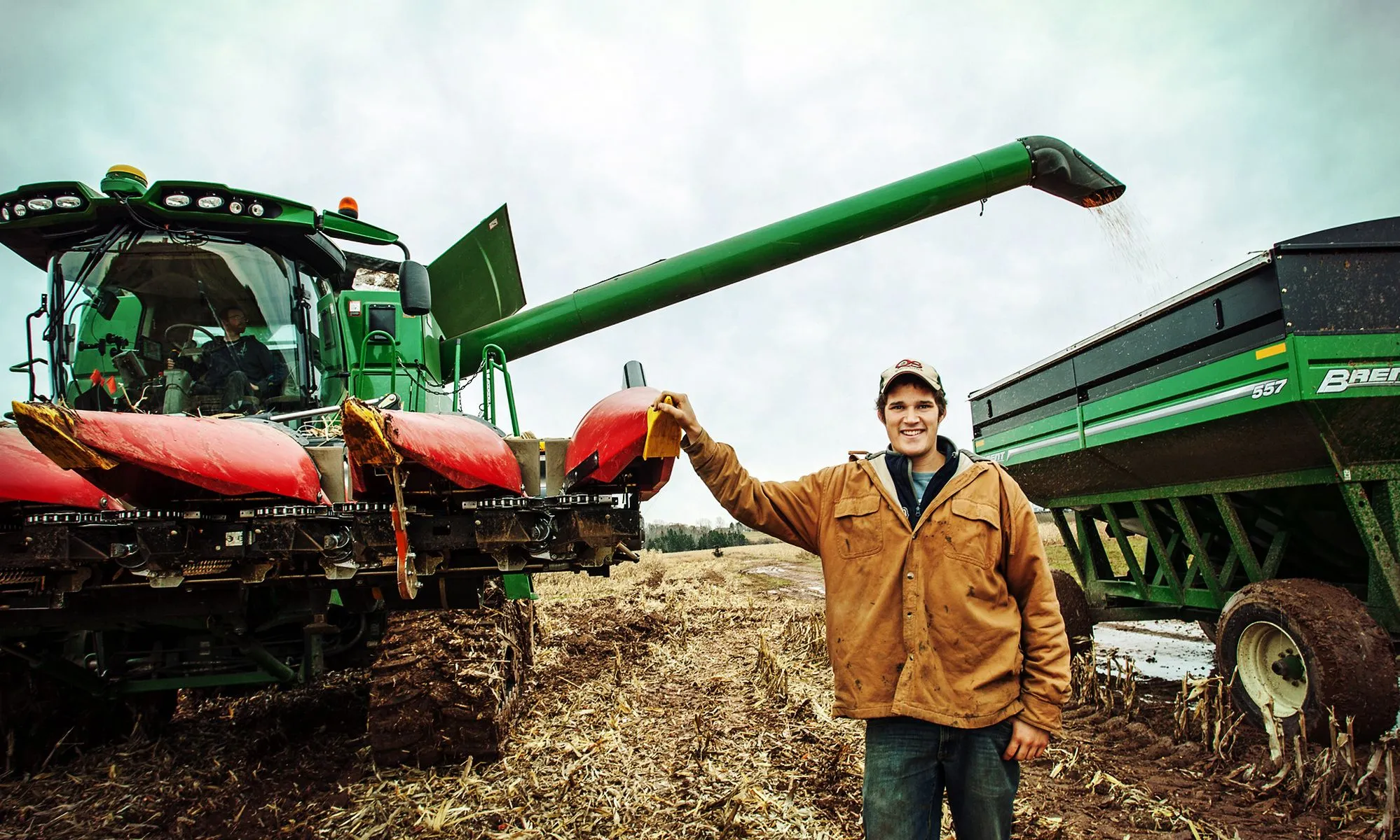 A smiling young farmer in a brown jacket stands in a harvested cornfield next to a large green and red combine and a grain cart with grain pouring into it.