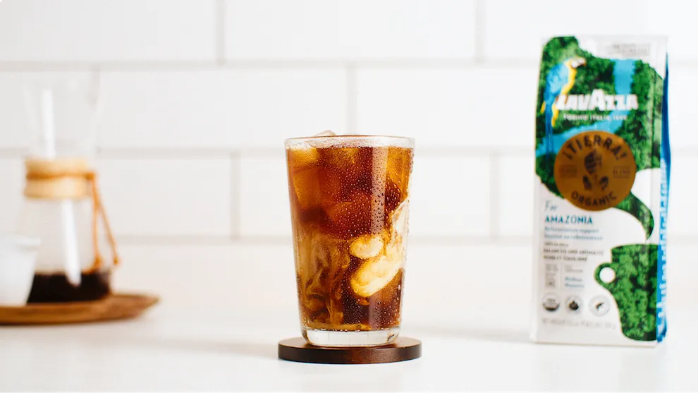 A tall glass of iced coffee with milk on a small wooden coaster, with a Chemex coffee maker in the background and a Lavazza Tierra! Organic coffee bag to the right, all on a white surface against a white tiled wall.