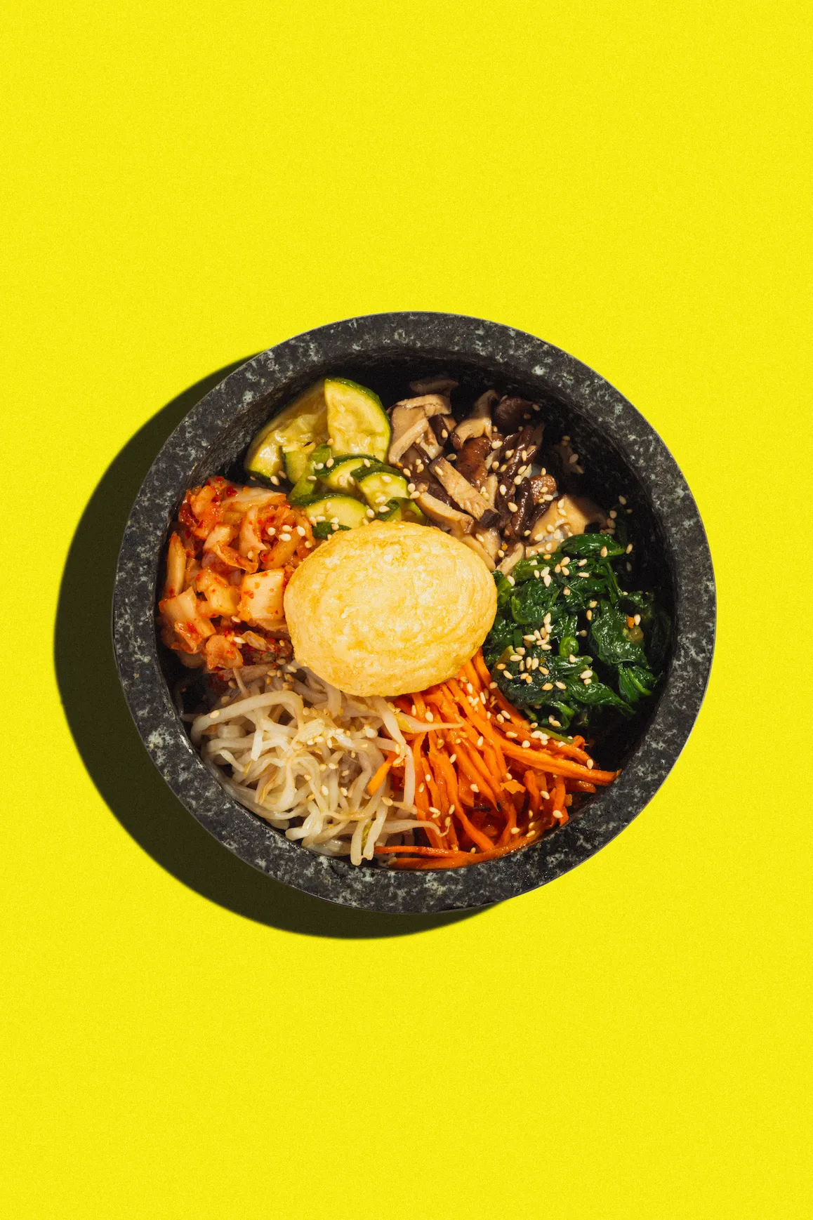 Overhead shot of bibimbap in a dark stone bowl on a bright yellow background, featuring various colorful vegetables and a fried egg.