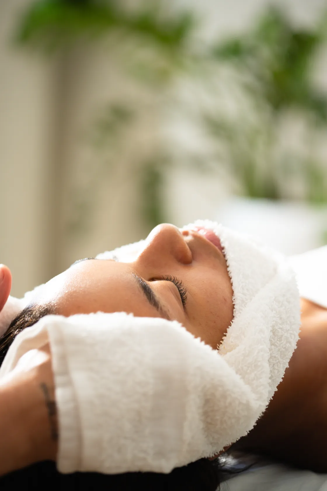 Close-up of a person lying down with a white towel wrapped around their head, eyes closed, with a green plant in the background.