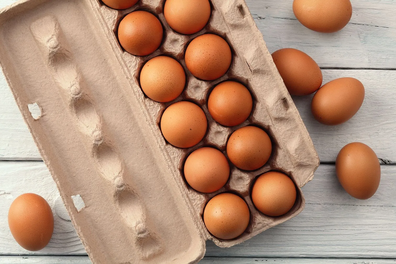 Overhead shot of brown eggs in a cardboard carton and loose on a light wooden surface.