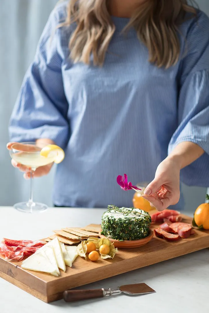 A woman with blonde hair holds a cocktail and a purple flower above a wooden charcuterie board with cheese, crackers, salami, and fruit.