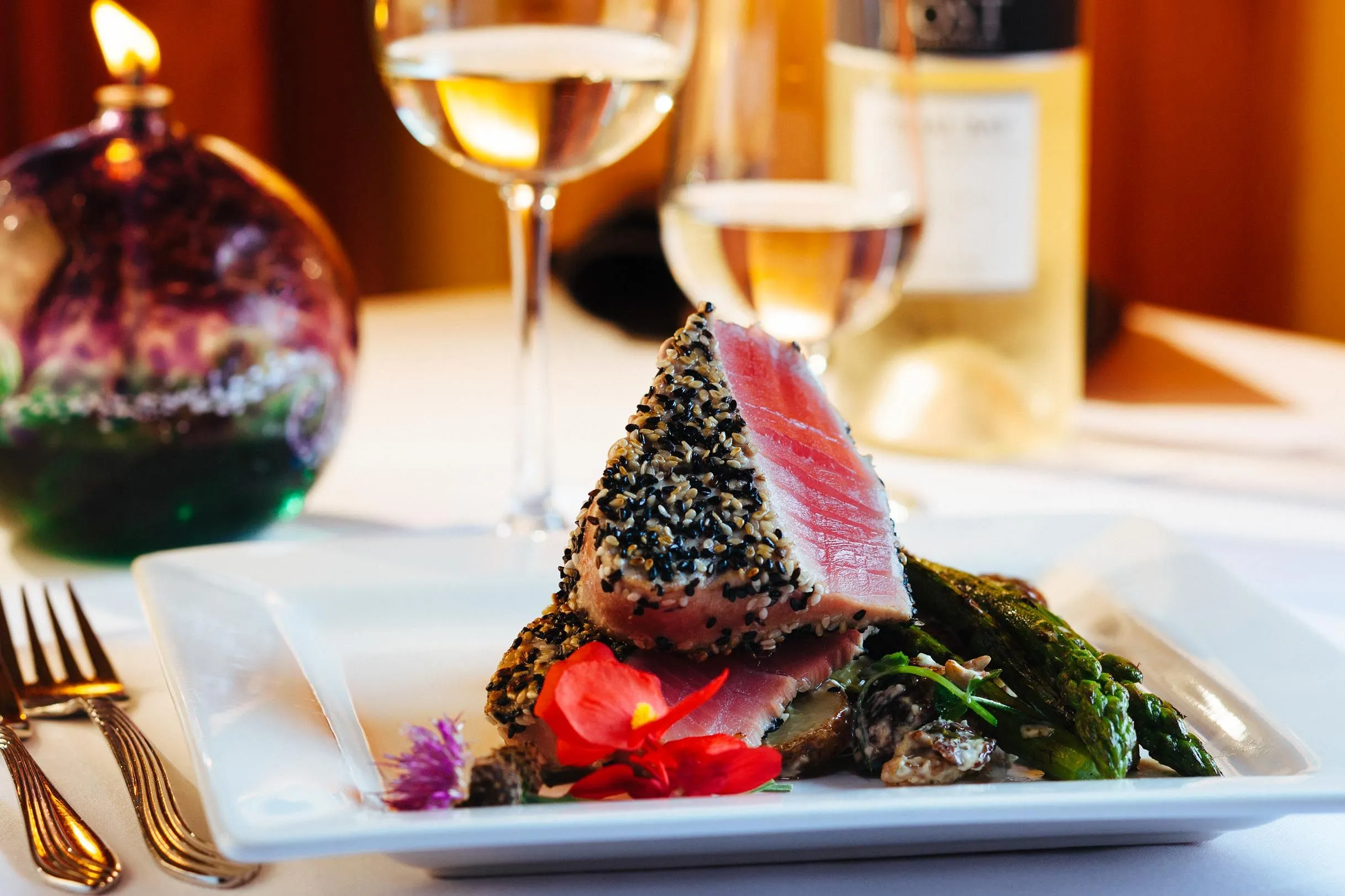 Close-up of a white plate with seared tuna, asparagus, and flowers; wine glasses are in the background.