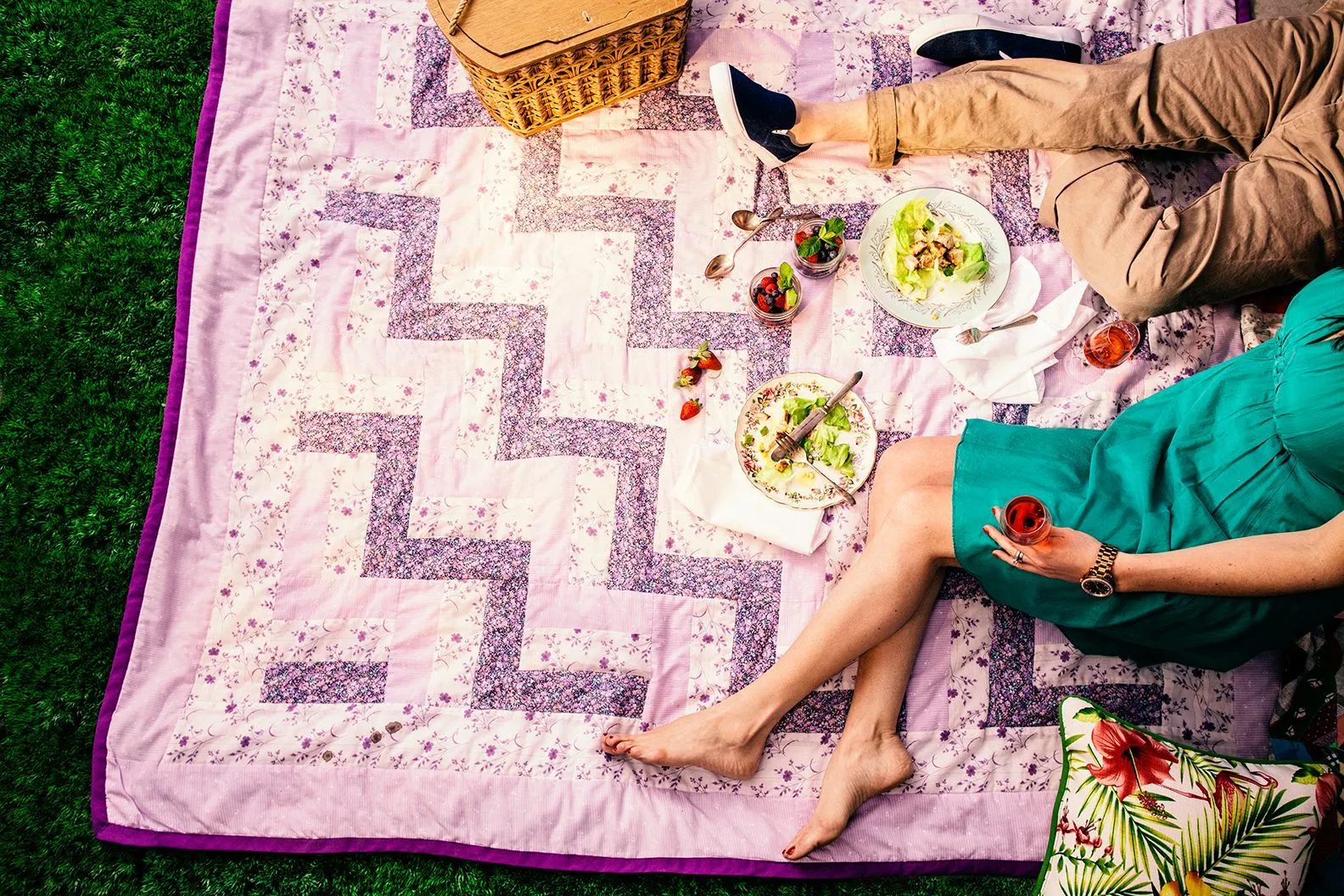 Overhead view of a picnic: two people relaxing on a patchwork quilt with salads, berries, drinks, and a picnic basket.
