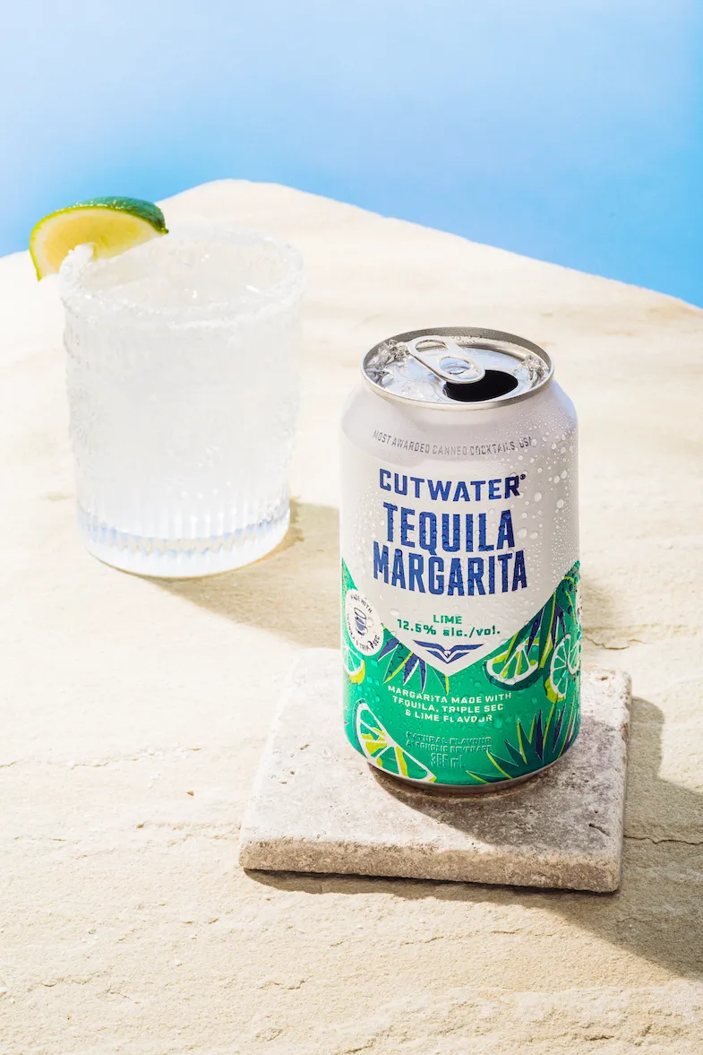 A can of Cutwater Tequila Margarita and a glass of margarita with a lime wedge, set against a light blue background and a beige textured surface.