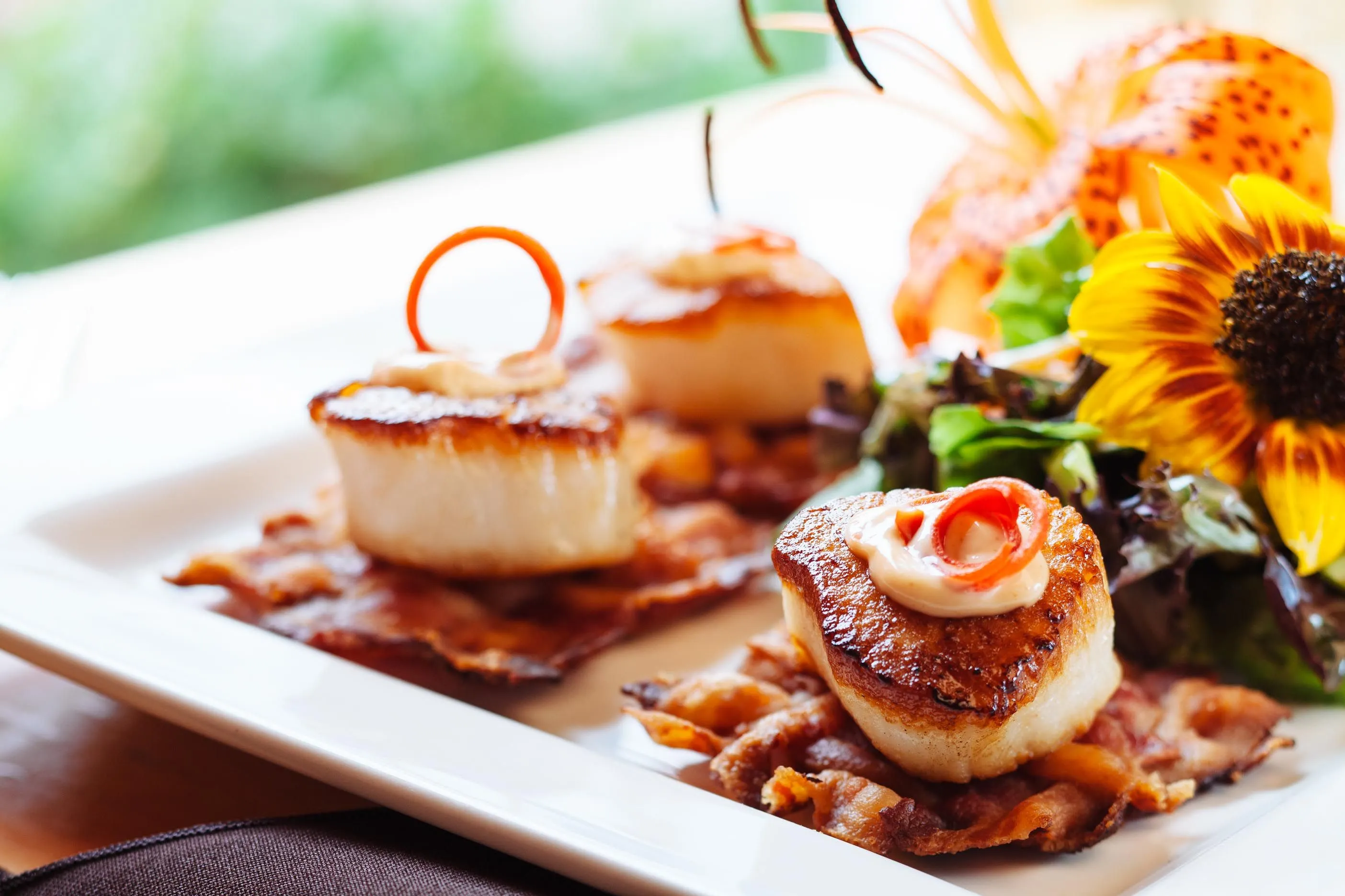 Three seared scallops with sauce and garnish atop bacon, served on a white plate with salad and flowers.