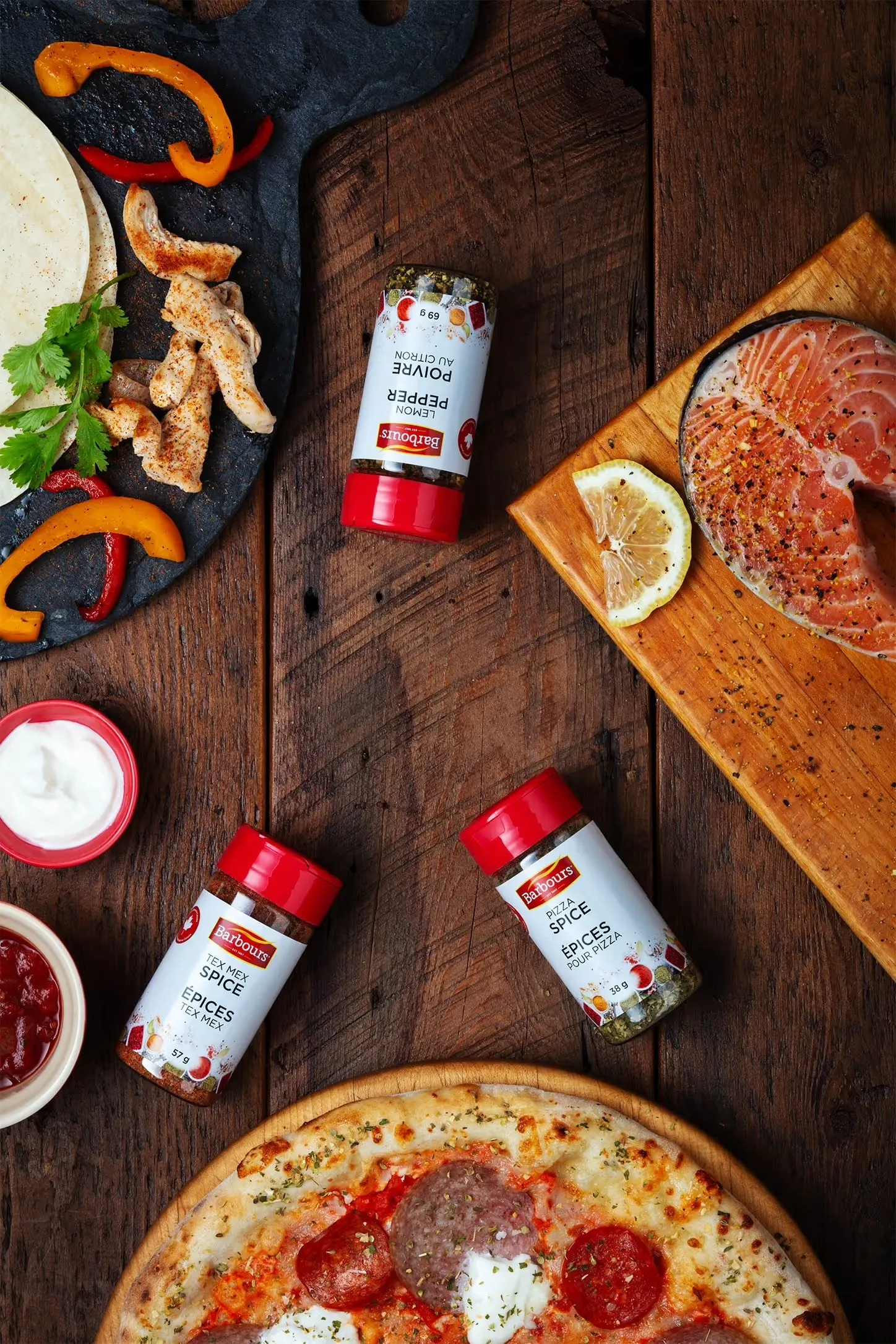 Overhead shot of a wood table with various food items including a pizza, salmon steak, tortillas, chicken, peppers, sauces, and spice jars.