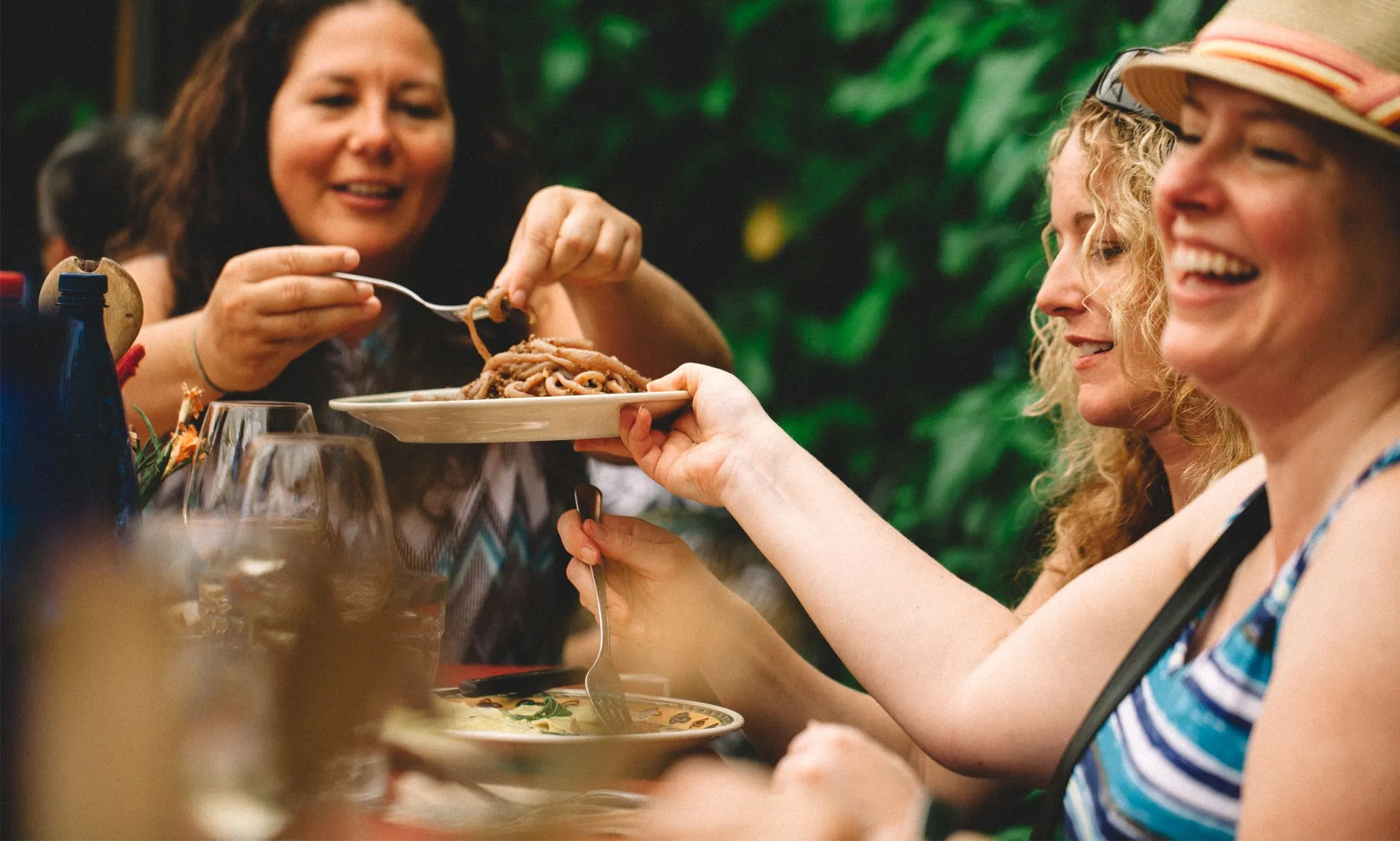 Several women enjoying an outdoor meal together, with one offering a plate of spaghetti to another, all smiling and surrounded by greenery.