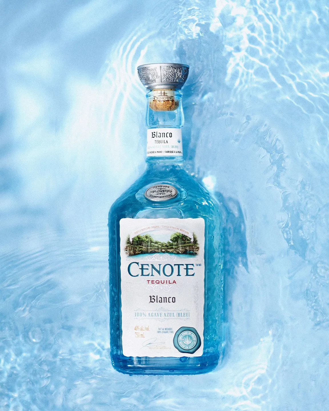 A blue glass bottle of Cenote Blanco tequila, with a silver cap and white label, submerged in shimmering blue water.